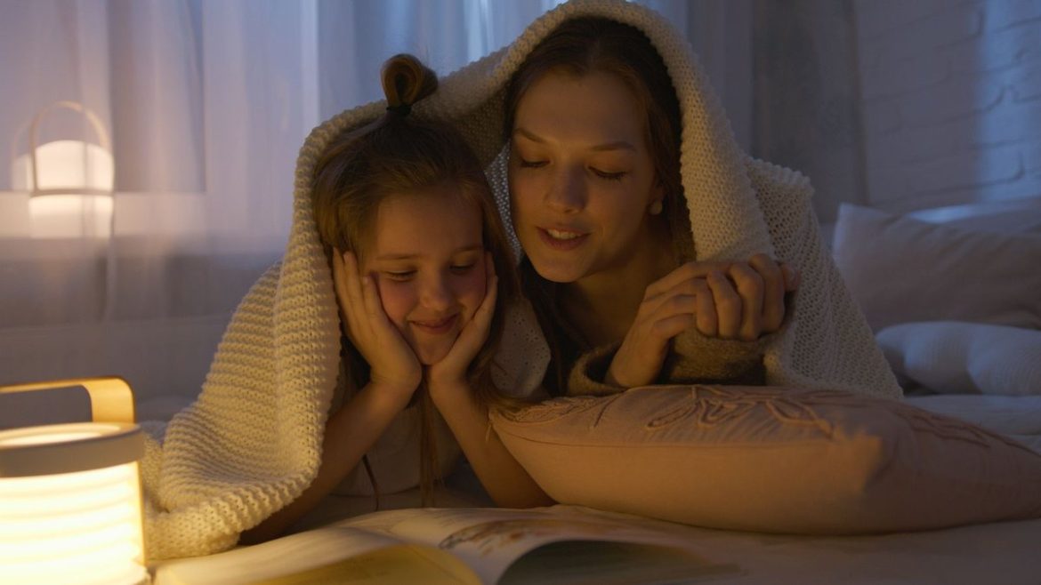 Мама з донькою читають книжку під ковдрою при теплому світлі